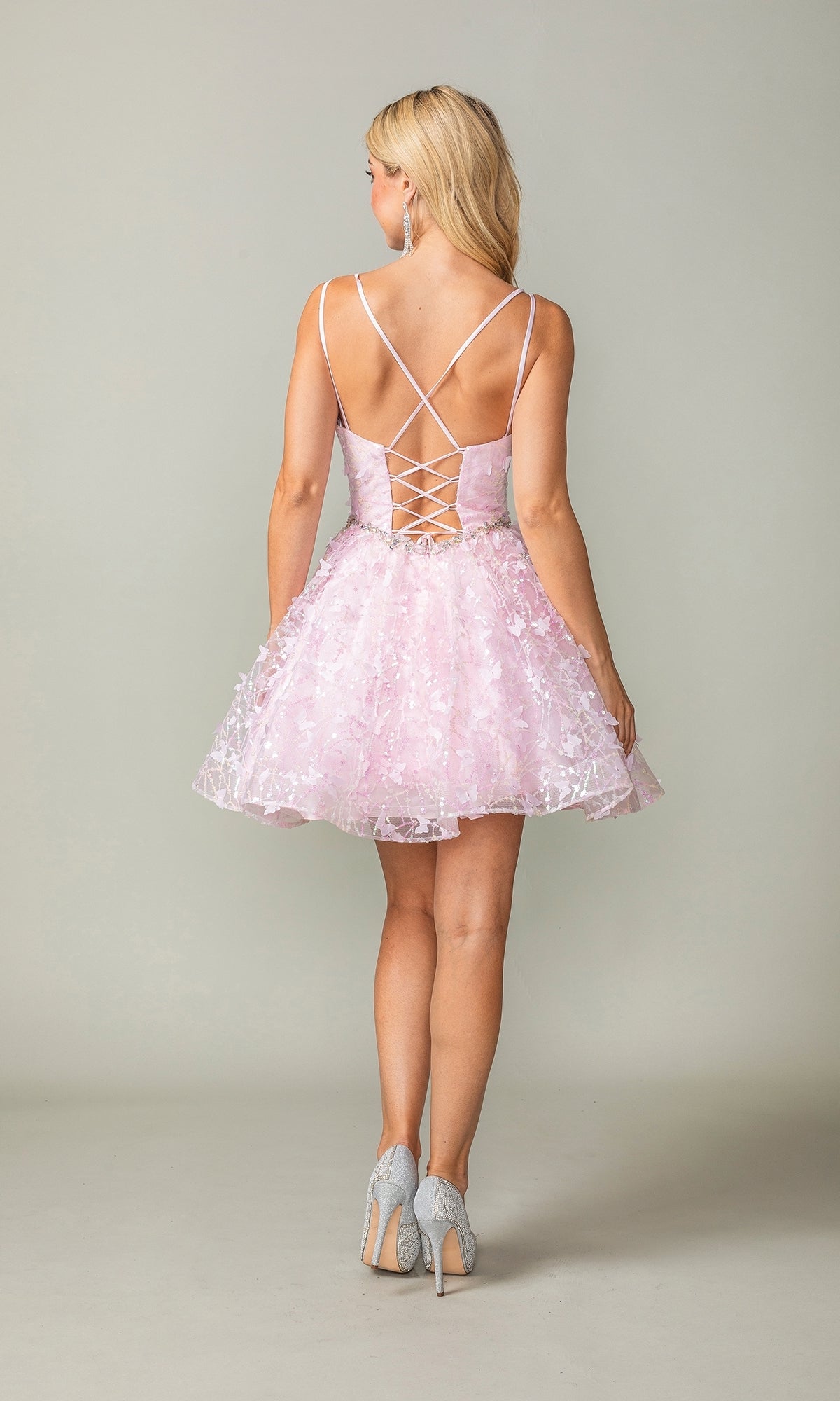 A woman with blonde hair faces away, wearing the Three-D Short Butterfly Homecoming Dress 3350 in light pink, featuring a lace-up back and corset-style bodice, paired with light-colored high heels.